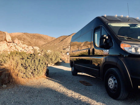 Black camper van RV rental parked on a desert road. Rocky mountains and clear sky reflected
