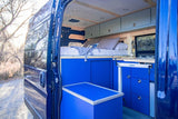 This RV rental camper van interior boasts a cozy bed, kitchenette, and bright blue cabinetry, ideal for mobile living.