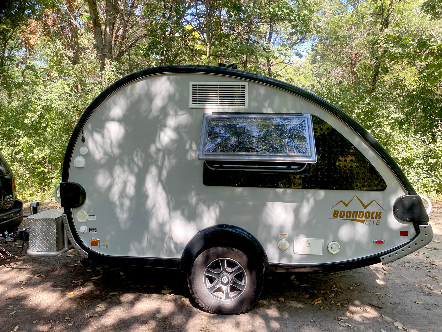 This light gray Boondock Lite teardrop RV rental is parked among trees, its side dappled with leaf shadows, ready for outdoor travel.