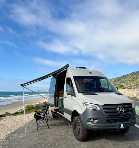 Adventure-ready Mercedes Sprinter camper van RV rental parked by the ocean with awning extended & chairs. Ideal beach getaway.
