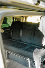 Interior of an RV rental: rear bench seat with seatbelts, window curtains, and wooden floor, ideal for camper van travel.