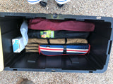 A large black storage bin for an RV rental is packed with camping gear: a folding chair, mat, electric shower, and other bags.