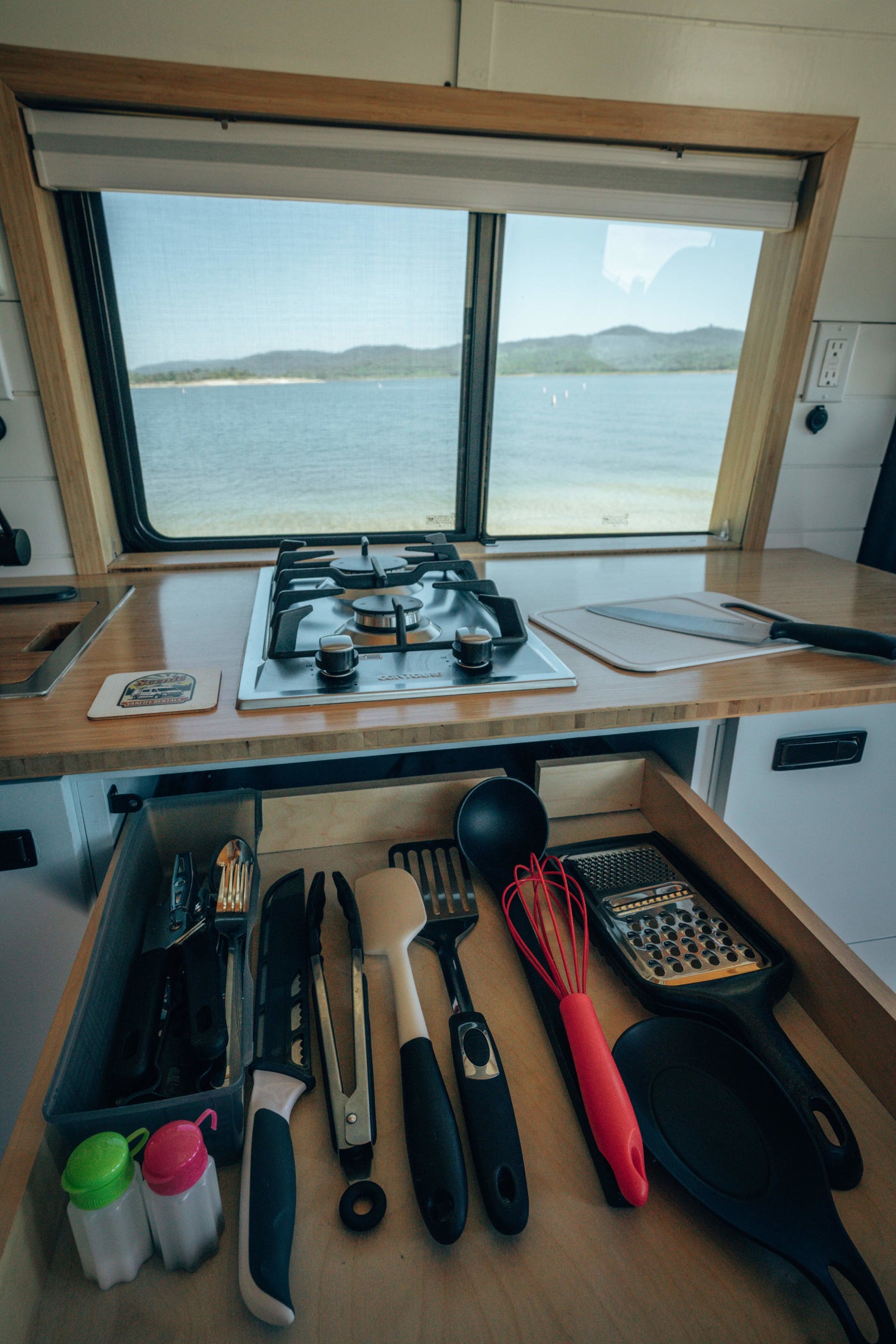 RV rental kitchen with a scenic lake view: built-in stove, counter, and a drawer stocked with cooking utensils.