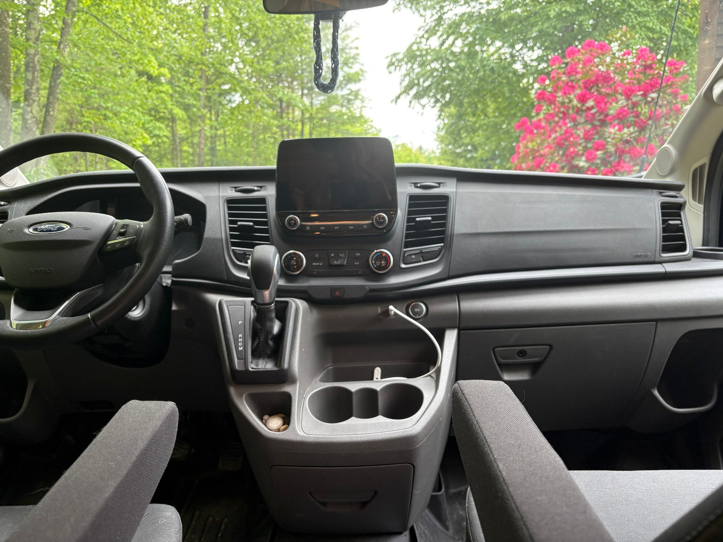 RV rental van interior: Ford dashboard, steering wheel, gear shifter, cupholders, with green trees & pink flowers visible outside.