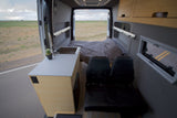 Inside this RV rental: a modern camper van interior with kitchen, bed, and seats. Open rear doors reveal a flat, open landscape.