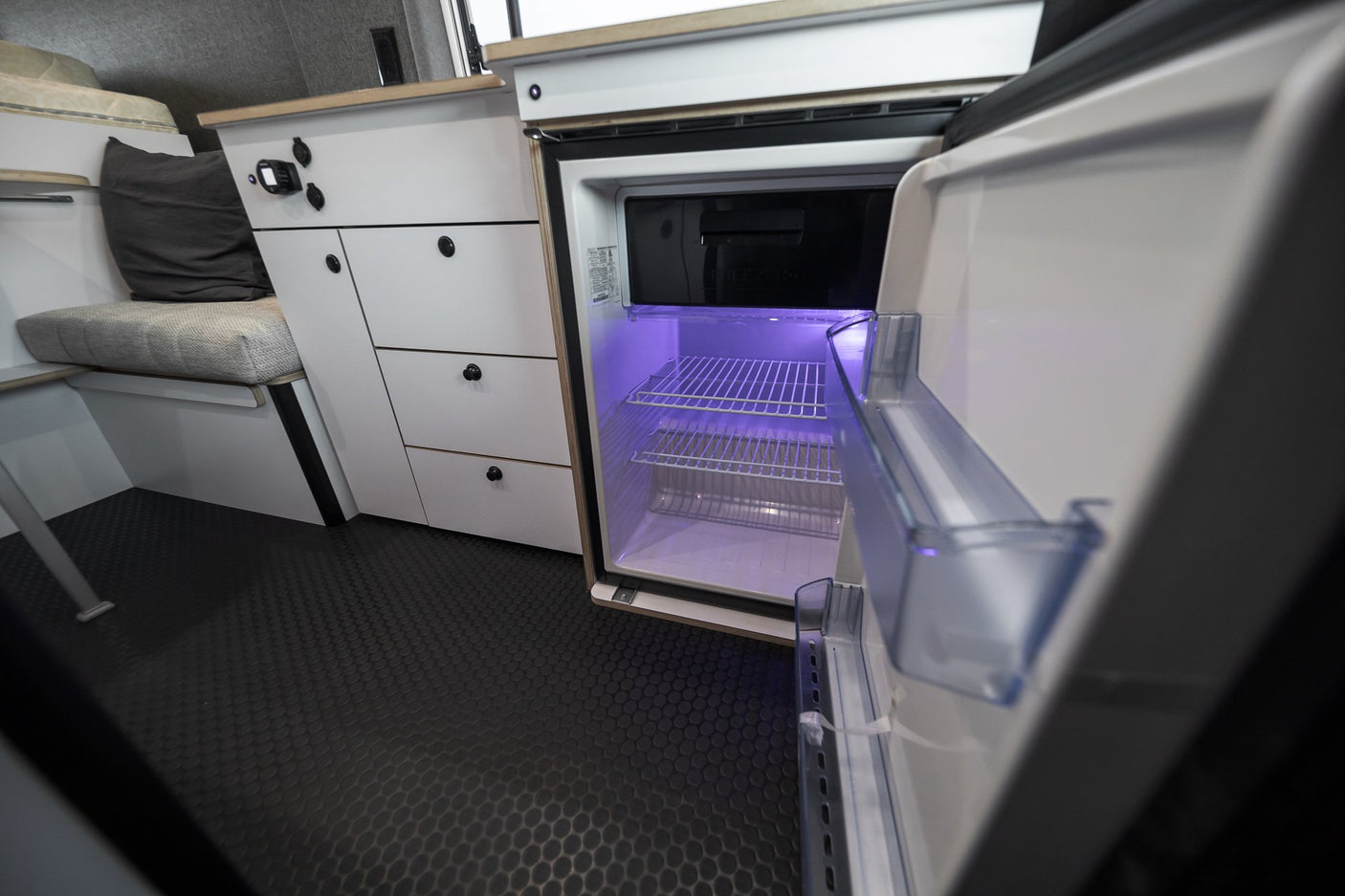 Modern RV rental interior: open fridge with purple light, white cabinets, and comfortable bench seating.