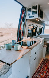 RV rental: Modern van kitchen with induction cooktop, coffee, and scenic field view from open back doors.