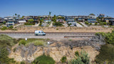 White & green RV rental on coastal road, train tracks, hillside homes, and dry cliffs under a clear sky.