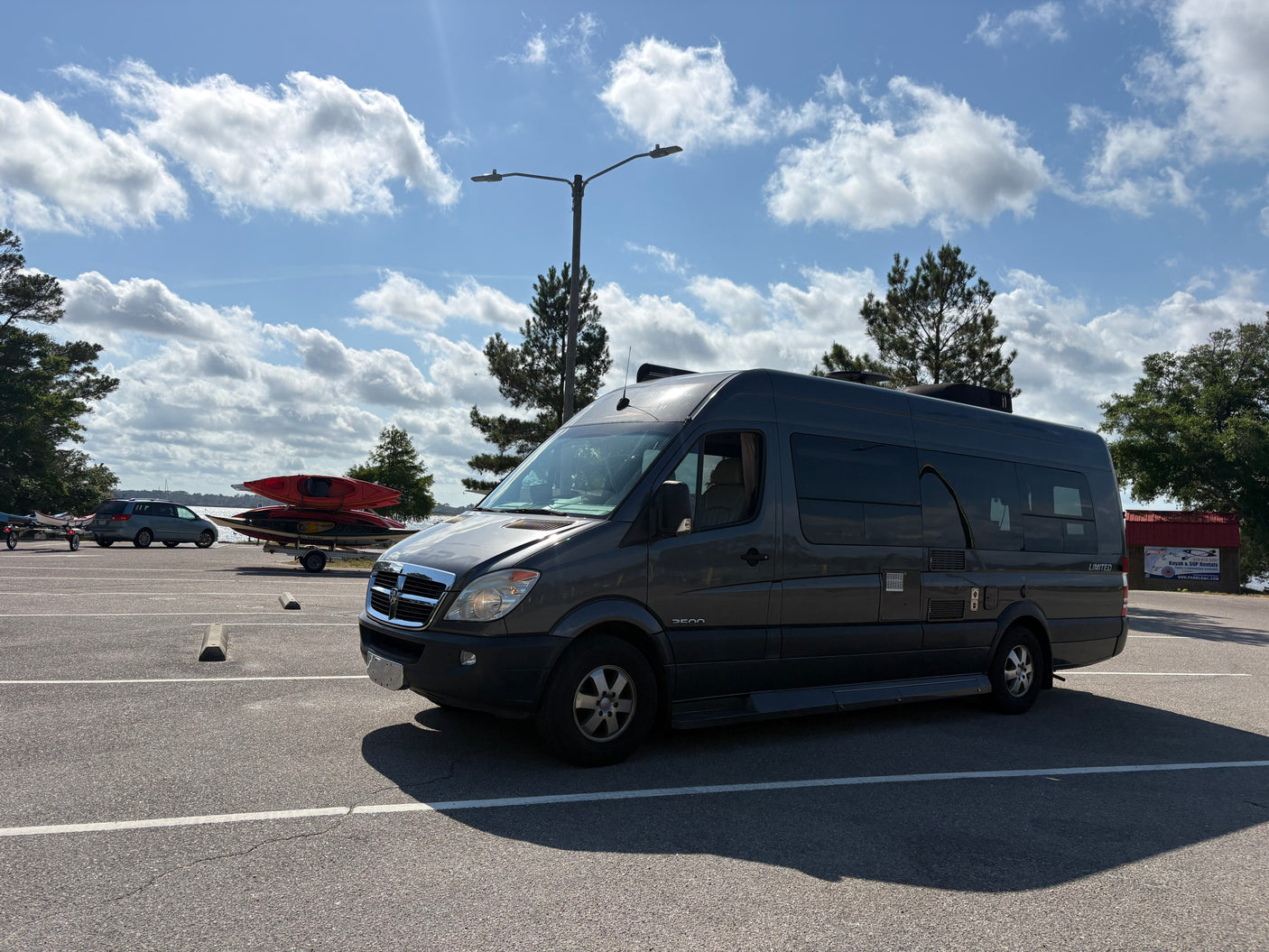 Grey RV rental van parked in a lot by a lake, with kayaks and a rental business in the background, ready for water adventures.