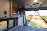 RV rental camper van interior showing a cozy bed, storage, and an open rear view of a sunny field through a bug net.