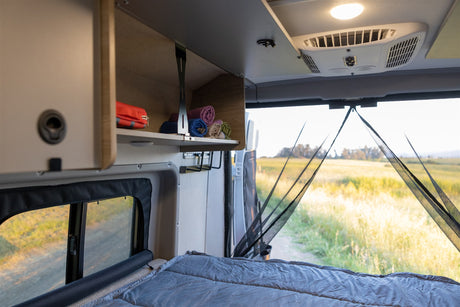 RV rental camper van interior showing a cozy bed, storage, and an open rear view of a sunny field through a bug net.