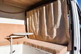 Inside an RV rental: a cozy living space with tan bench seating, a white adjustable table, and a light privacy curtain.