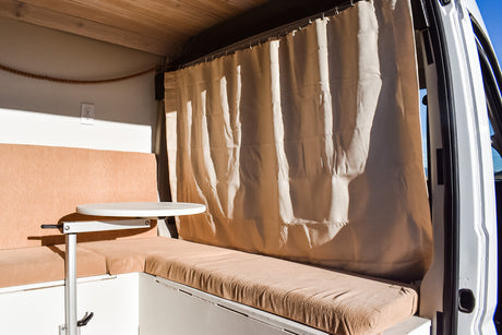 Inside an RV rental: a cozy living space with tan bench seating, a white adjustable table, and a light privacy curtain.