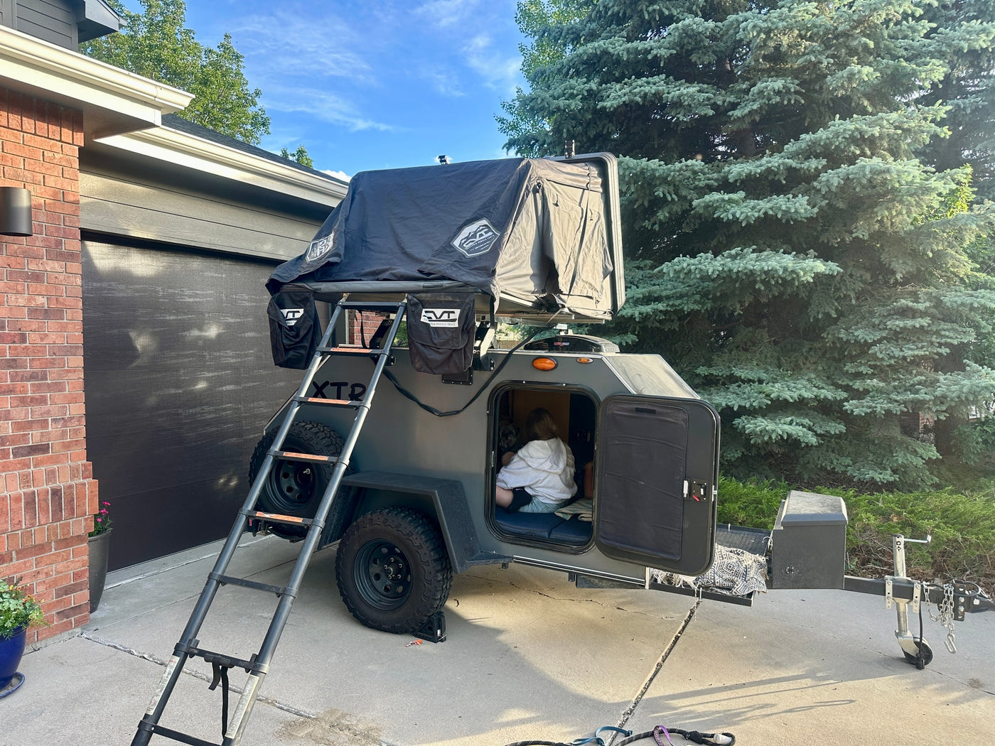 RV rental: Grey teardrop camper trailer with roof-top tent and ladder, parked. A person is comfortably inside. — photo 1
