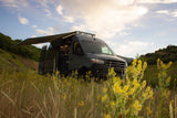 Adventure awaits! Dark gray Sprinter RV rental with awning extended, parked in a field of yellow flowers.