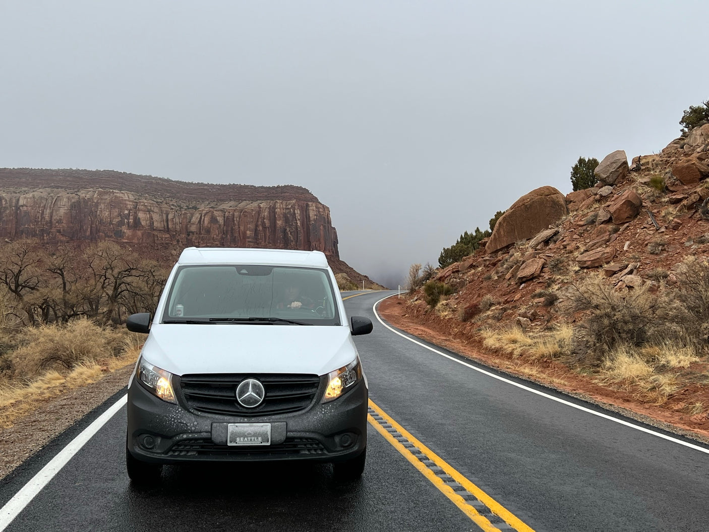A white Mercedes camper van, an RV rental, on a wet road in a dramatic red rock canyon under an overcast sky.