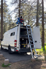 A person relaxes on the roof of a white RV rental camper van, parked in a sunny forest. A ladder extends from the open back doors.