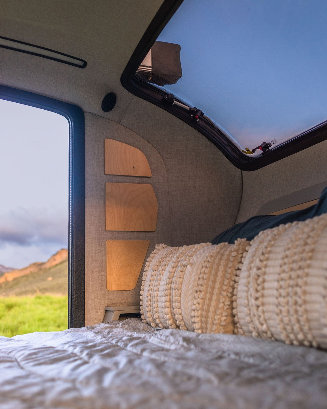 Inside a cozy RV rental: a comfortable bed, wooden shelves, and a large skylight showing blue sky, with a scenic outdoor view.