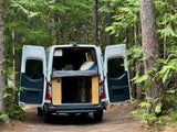 White camper van (RV rental) with rear doors open, revealing a cozy bed, parked on a forest path for outdoor adventures.