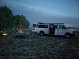 An RV rental provides evening calm for a family camping trip. A white van is parked next to a warm campfire, with a child enjoying the starry night outdoors.