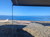Enjoying a stunning ocean view from an RV rental on a sunny pebble beach with driftwood, distant land visible across the water.