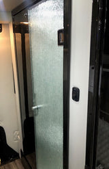 Modern compact shower in an RV rental, featuring a textured glass door with a silver handle and an adjacent light switch.