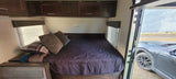 Cozy RV rental bedroom with dark bedding, overhead cabinets, and window blinds. A car is visible outside.
