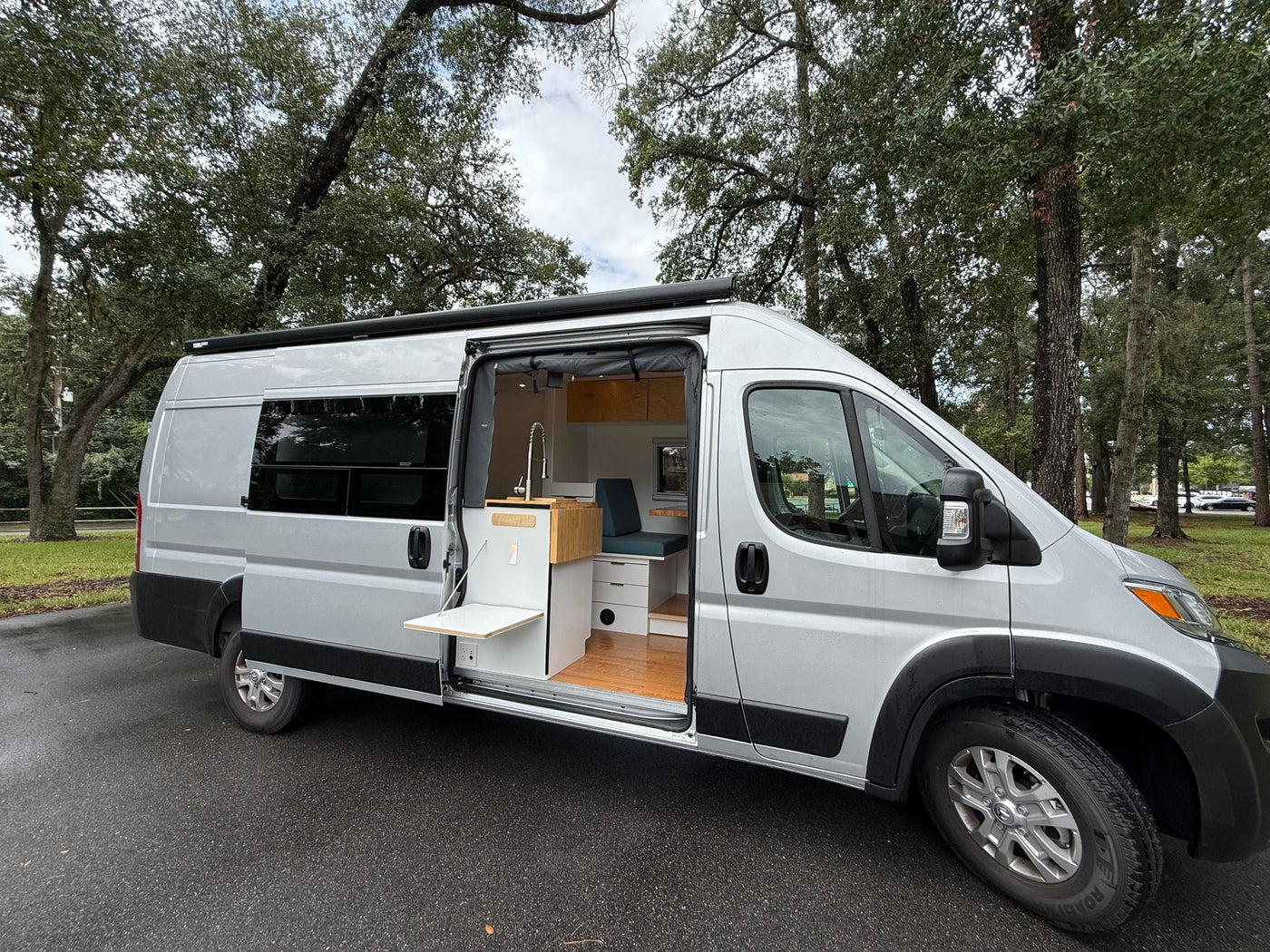  parked by green trees under an overcast sky.|RV rental van interior features a comfy bed — photo 15