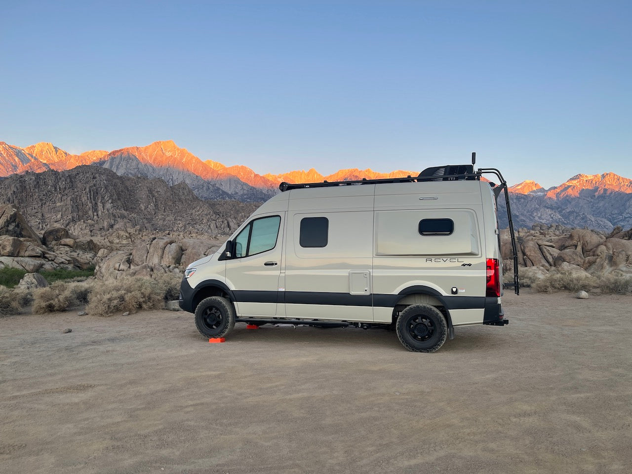 Scenic RV rental parked in a vast, rocky desert. Majestic mountains glow orange from the rising or setting sun behind it.