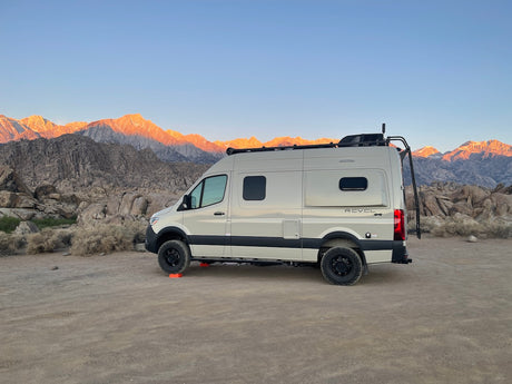 Scenic RV rental parked in a vast, rocky desert. Majestic mountains glow orange from the rising or setting sun behind it.