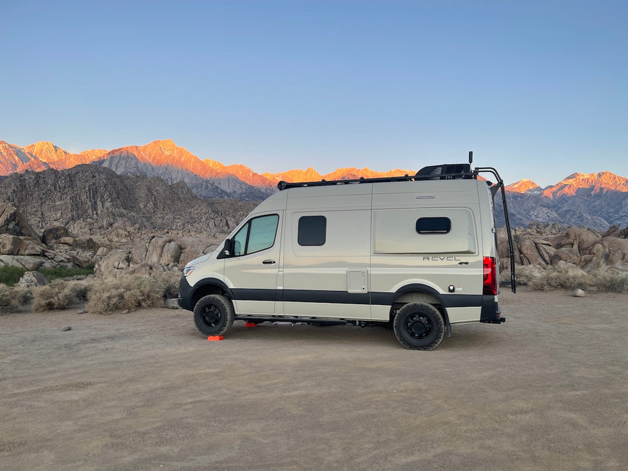 Scenic RV rental parked in a vast, rocky desert. Majestic mountains glow orange from the rising or setting sun behind it.