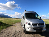 A silver Mercedes Sprinter RV rental van on a scenic gravel road, with snow-capped mountains & blue sky. Ideal for adventure.