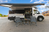 Ethos RV rental: silver camper van w/ extended awning, chairs & table by a scenic river & mountains.