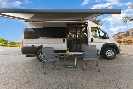 Ethos RV rental: silver camper van w/ extended awning, chairs & table by a scenic river & mountains.