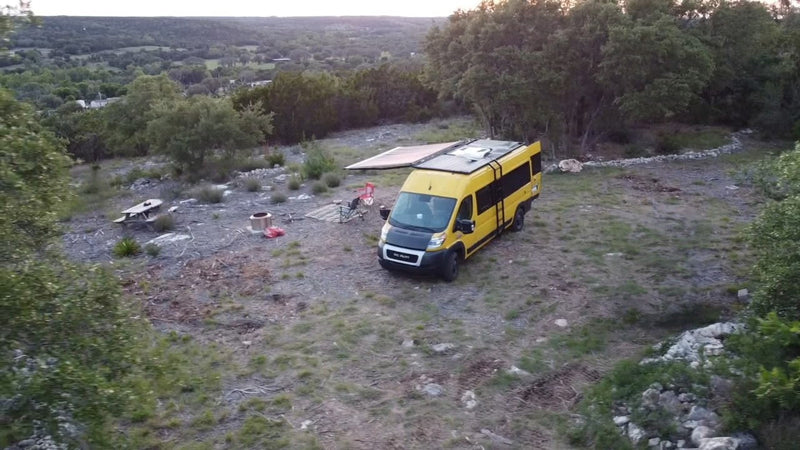 Yellow RV rental camper van in a scenic natural setting. Awning extended, solar panels, picnic table, and fire pit visible. — photo 8