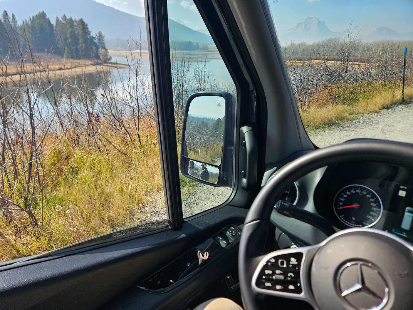 RV rental view of serene lake, mountains, and dirt road from inside the van. Side mirror shows two people.