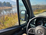 RV rental view of serene lake, mountains, and dirt road from inside the van. Side mirror shows two people.