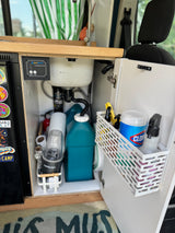 RV rental's under-sink utility area with LevelGuard tank monitor, water storage, blender, and cleaning essentials in a basket.