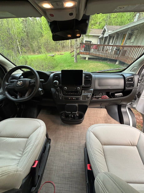 Inside this RV rental: front cabin with comfortable seats, a modern dashboard, and a scenic forest view outside a home.