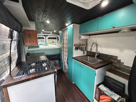RV rental: Custom van interior with functional kitchen, teal cabinets, cozy bed, and rustic wood finishes for van life.