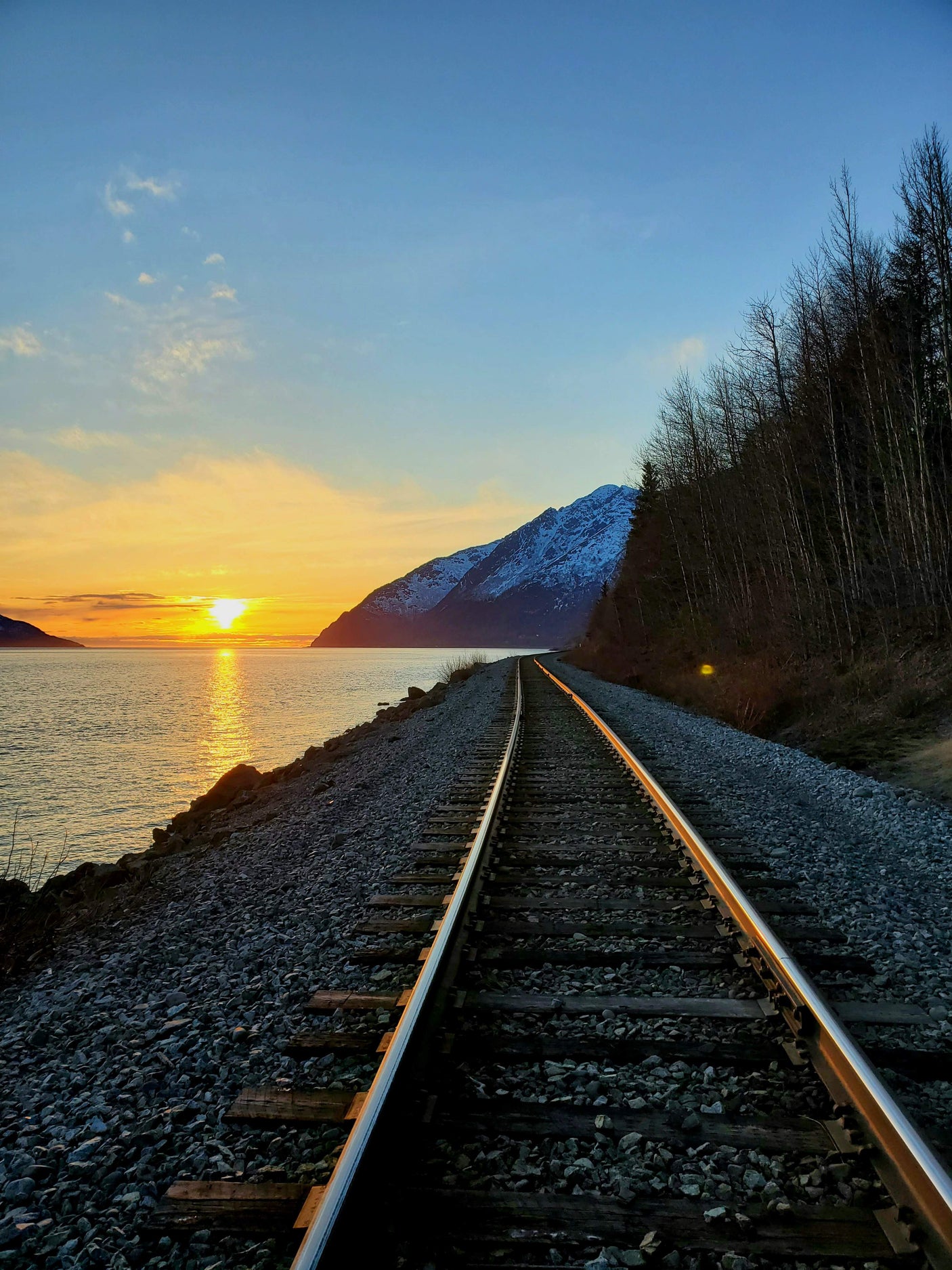 A vibrant sunset glows over water as railroad tracks curve past a snow-capped mountain and bare trees. Explore such views with an RV rental.