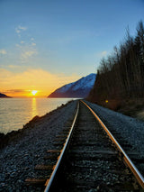 A vibrant sunset glows over water as railroad tracks curve past a snow-capped mountain and bare trees. Explore such views with an RV rental.