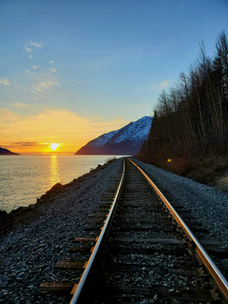 A vibrant sunset glows over water as railroad tracks curve past a snow-capped mountain and bare trees. Explore such views with an RV rental.