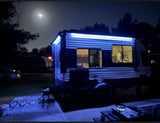 Camping at night in an RV rental. A blue-lit travel trailer shows a person inside, with an outdoor rug and grill setup under the moon.
