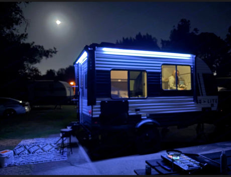 Camping at night in an RV rental. A blue-lit travel trailer shows a person inside, with an outdoor rug and grill setup under the moon.