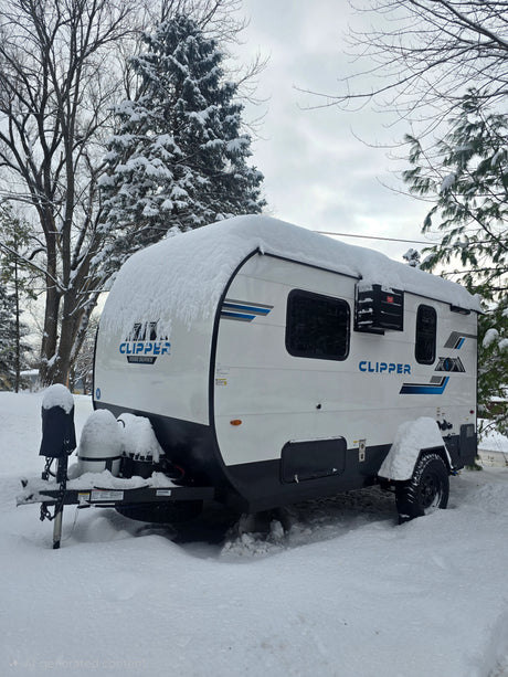 A white Clipper travel trailer, an RV rental, heavily covered in fresh snow, parked in a wintry landscape with bare and evergreen trees.