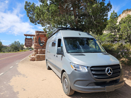 Grey rv rental van at Zion National Park entrance, with the park sign, road, and cliffs in view.