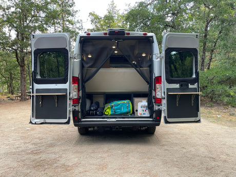 Open back of an RV rental camper van shows interior bed, storage, and fold-out tables for camping.