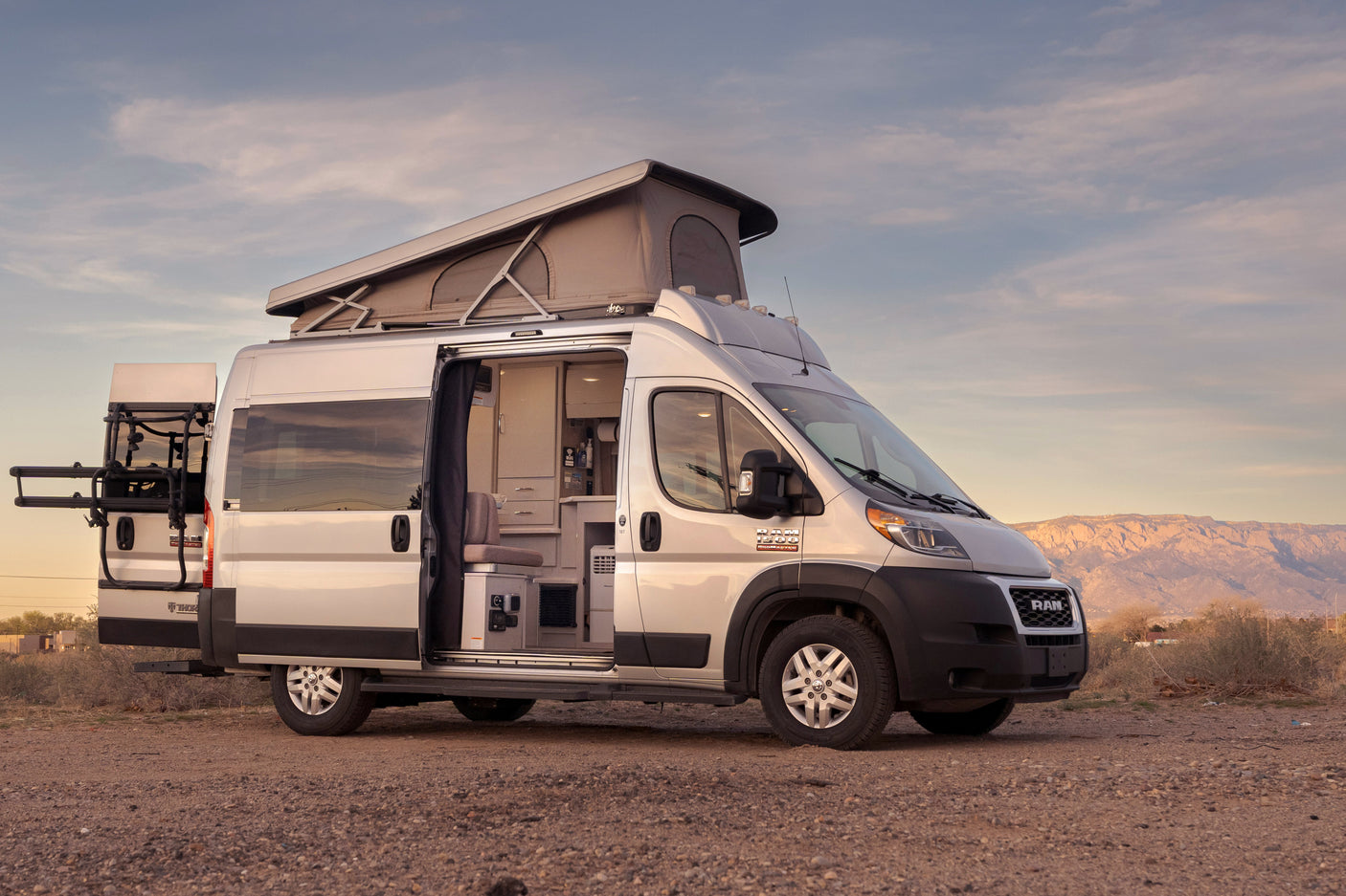 Silver pop-top RV rental in a desert landscape with mountains. Open doors reveal the camper van's inviting interior.