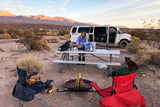 RV rental desert camping: Woman cooking at camp beside her van with a dog & campfire, mountains under sunset glow.
